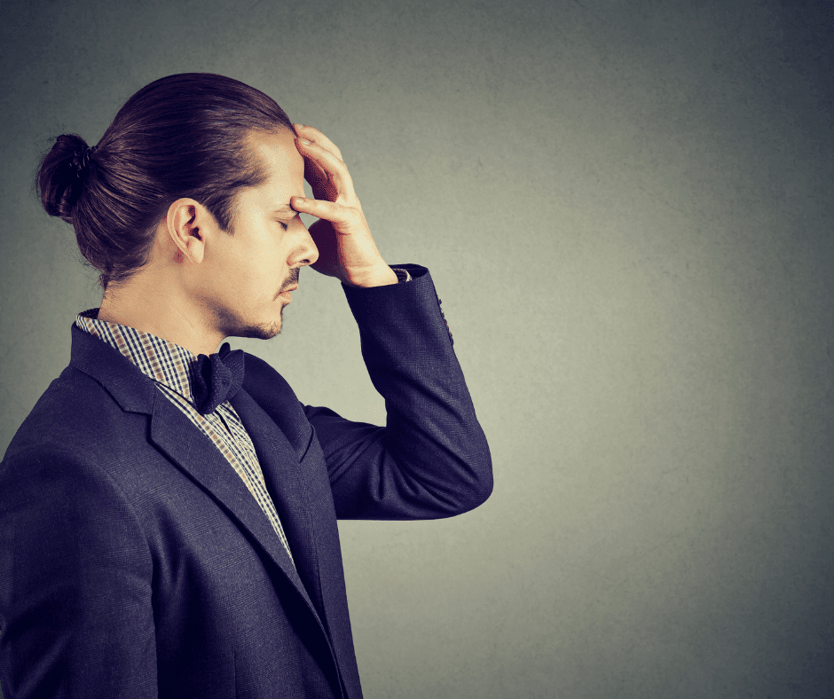 Person with light skin and dark hair pulled back into a bun and wearing a suit places his head in his hand with a concerned look on his face