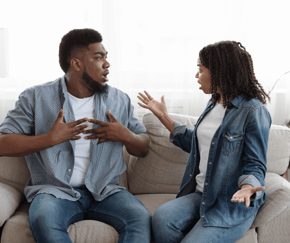 A couple, both with dark skin wearing denim, are sitting on a couch. They are clearly in conflict with one another.