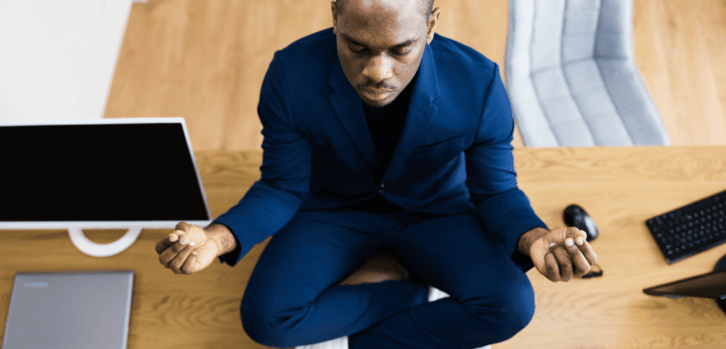 A black male sits on the floor next to his computer in a meditation stance