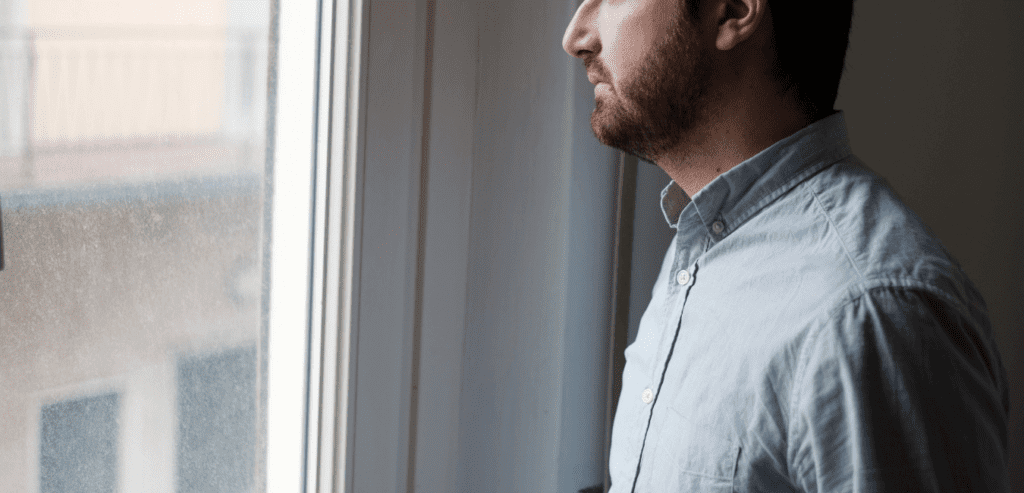 Man with beard wearing blue polo looking unhappy outside window