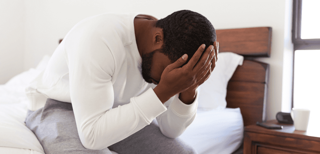 African man sitting on bed with his hand on head looking unhappy