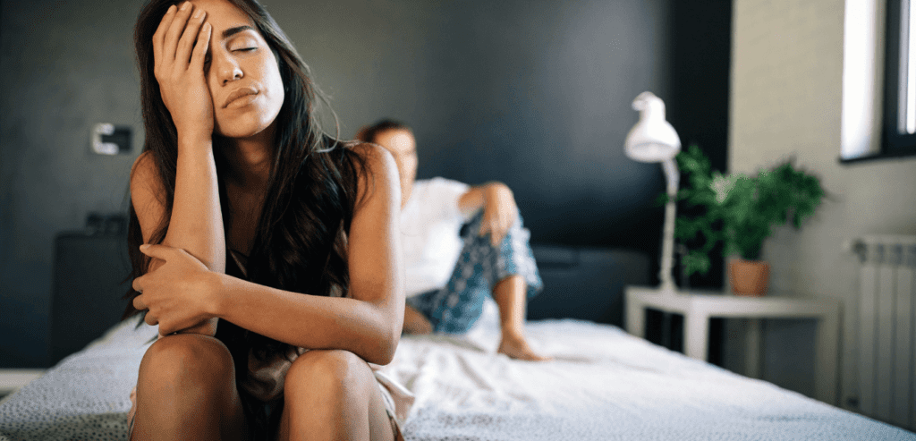 An upset female with her hands on her face sitting next to her partner on a bed 