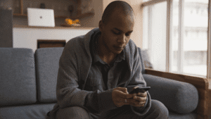 Man of color sitting on a sofa at home, holding a phone with a serious expression and his hand on his head.