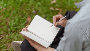 Close-up shot of a man writing in a notebook while sitting in a field.