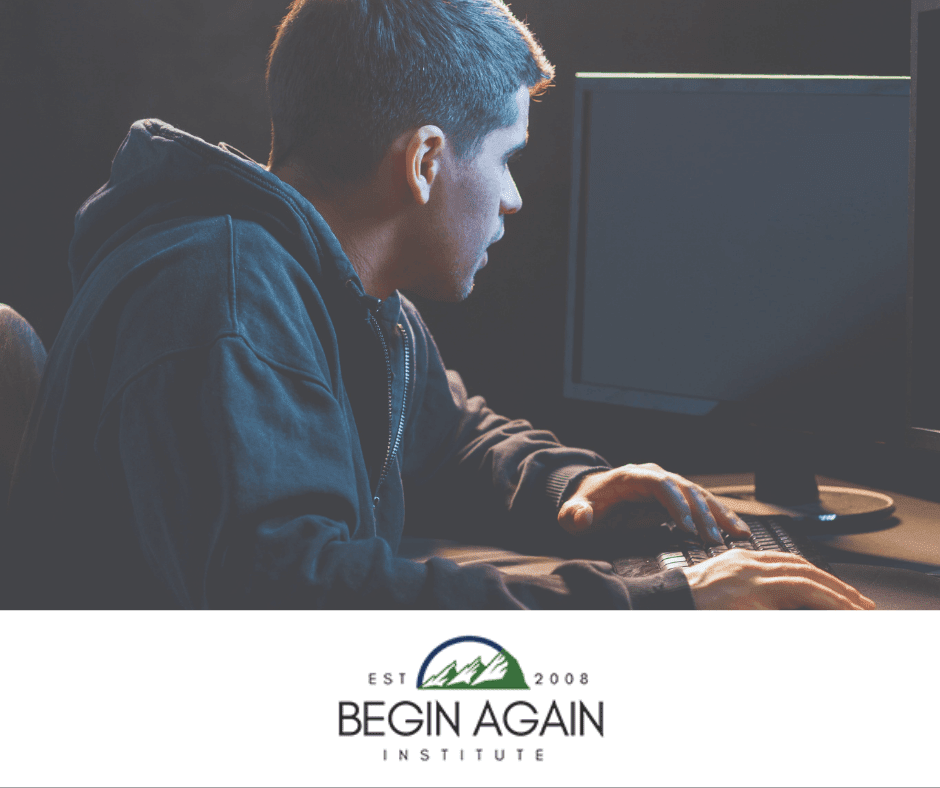 A person who identifies as male powers up a computer. The room is dark and he is wearing a hoodie.