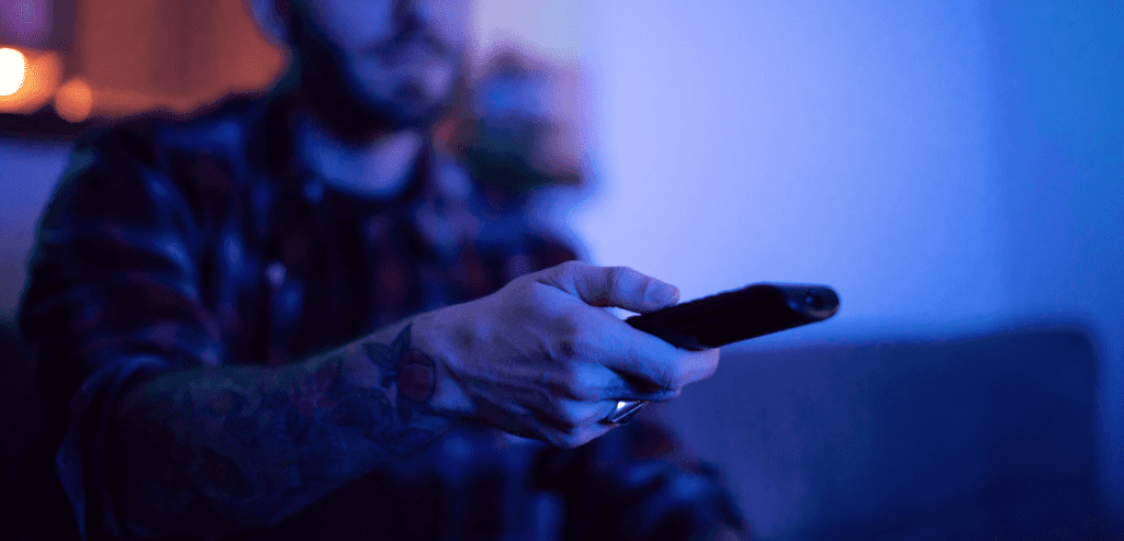 Man holding a remote in a dark room watching sexual pleasure videos as an escape