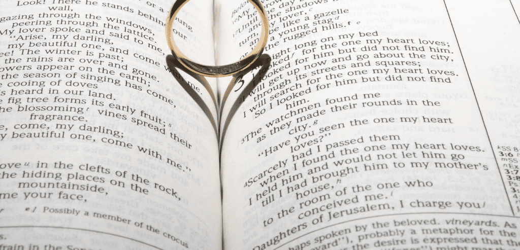 A wedding ring on a bible open to marriage scripture having a heart shaped shadow on it