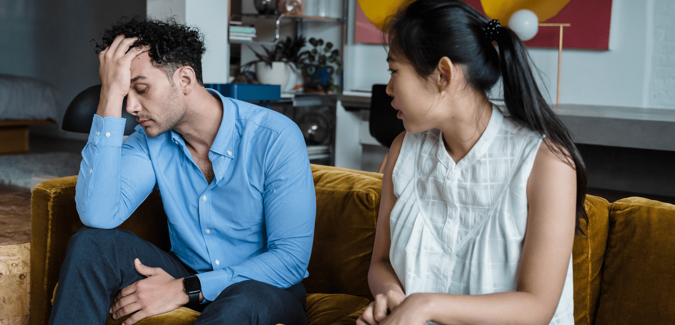A man and woman sit on a couch and are looking upset, he is holding his head in one hand