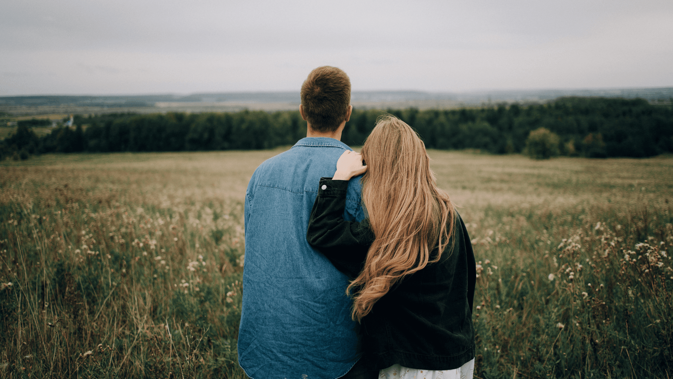 Back view of a couple standing outdoors, looking at green fields, with the woman’s hands resting on the man’s shoulders.