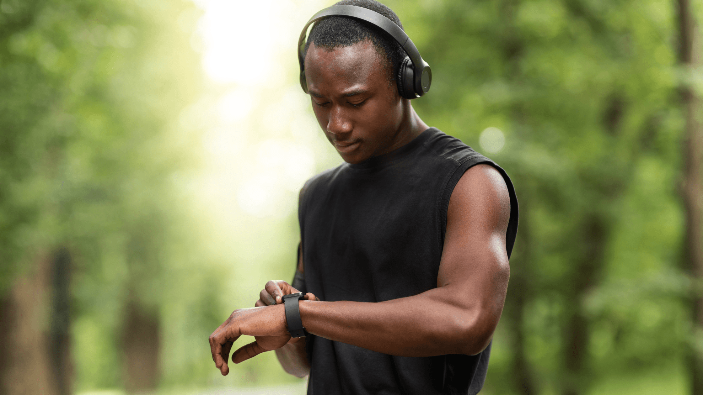 Man of color outdoors in headphones, looking at his smartwatch before running.
