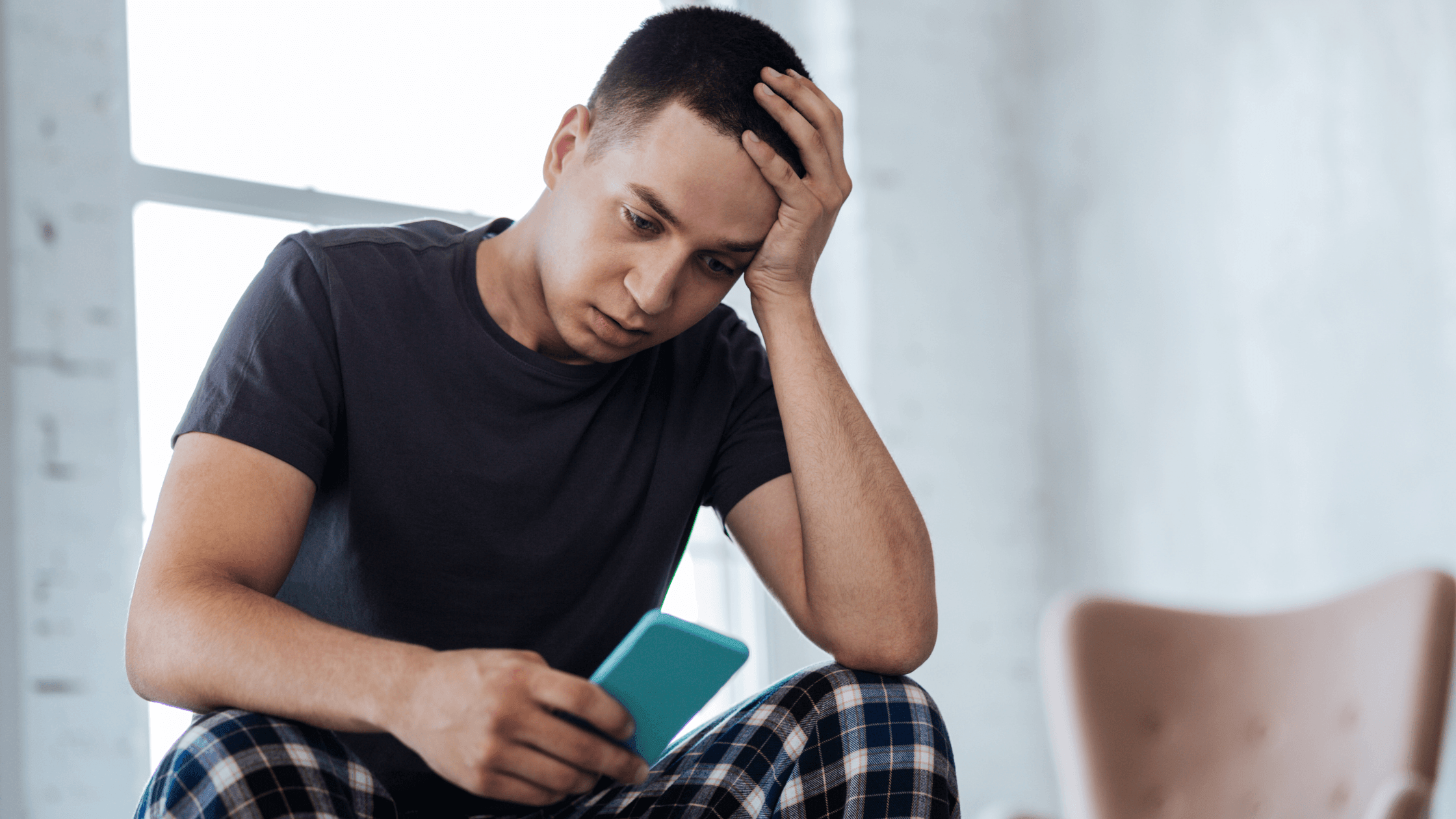 Man sitting on a couch in sleepwear, holding a phone with his hand on his head, looking stressed.