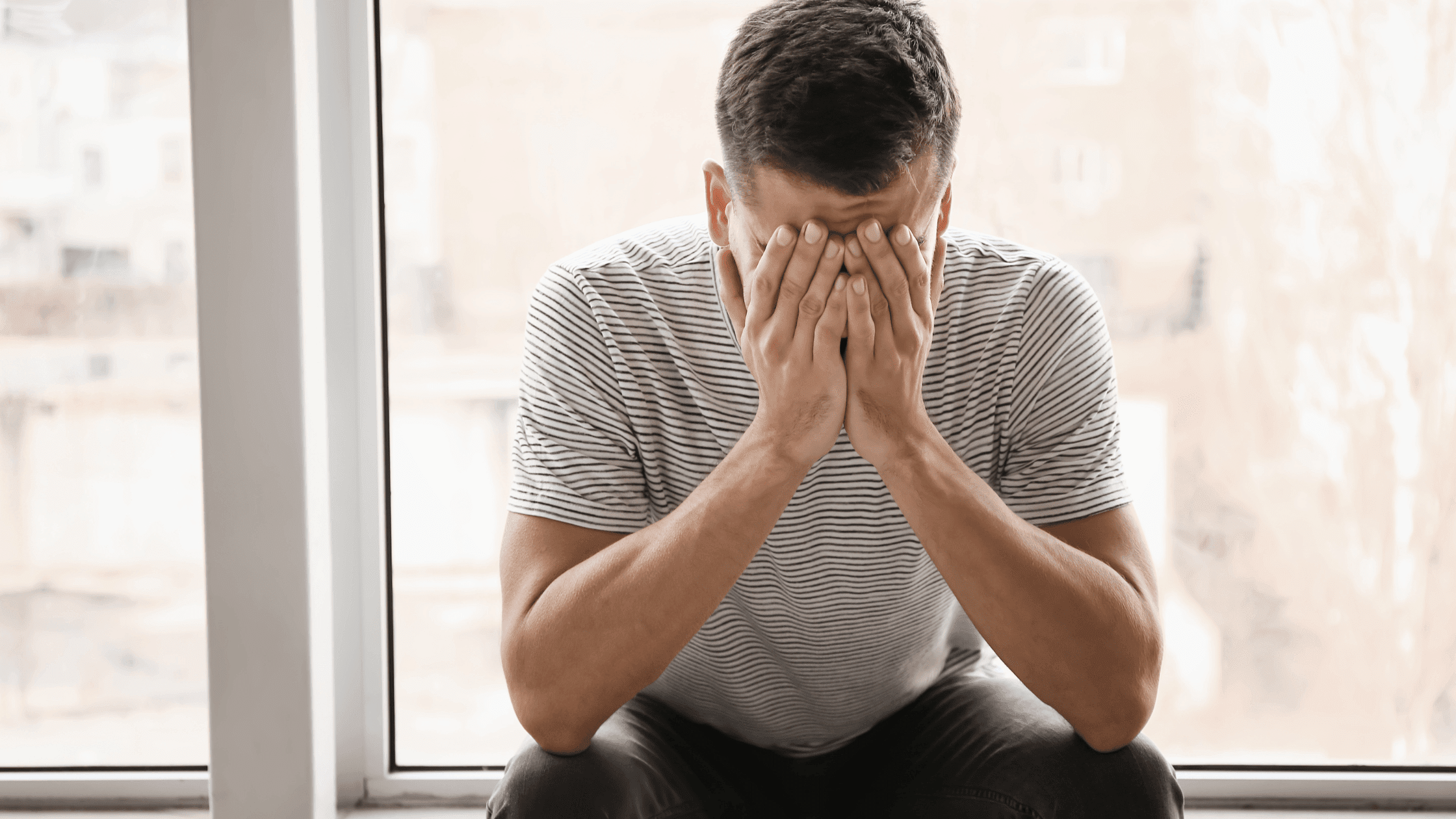 Man alone at home near windows, looking unhappy with his head lowered and hand on his face.