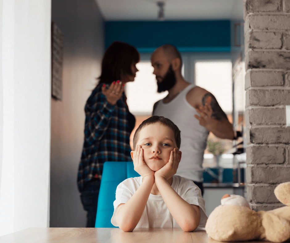 A child stares blankly ahead while his parents argue in the background. His father's hand is raised.