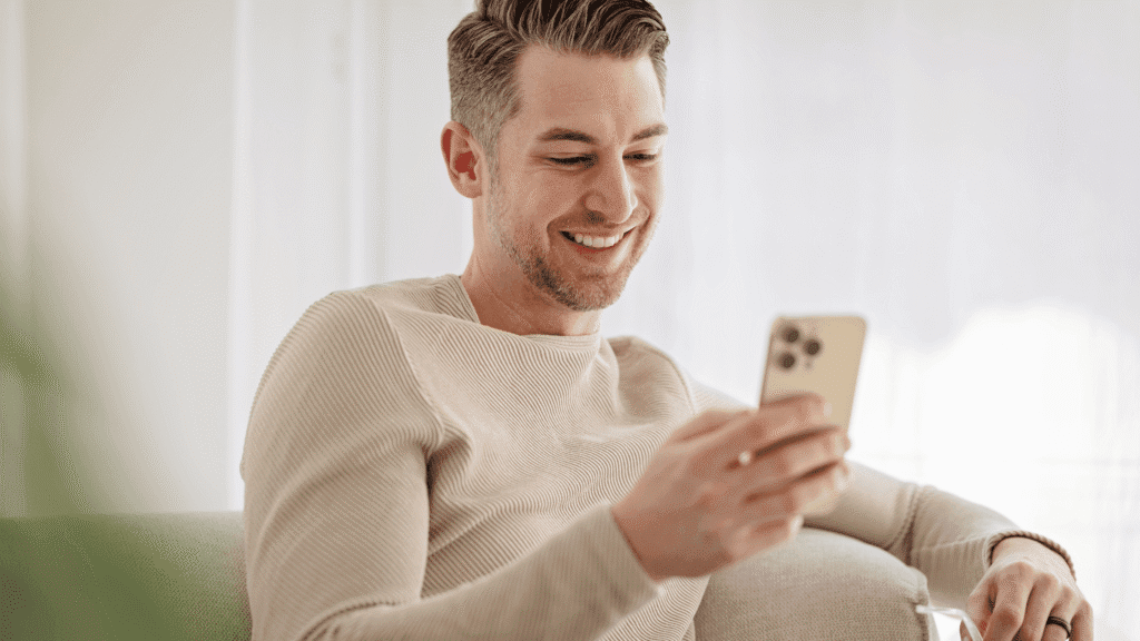 Man sitting comfortably on a couch at home, wearing a sweater, smiling while using a phone.