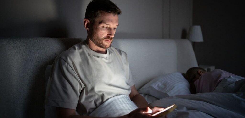 Man using smartphone in a dark bedroom while his wife sleeps beside him.
