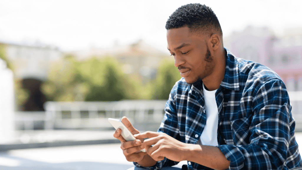 Man of color sitting outdoors on a sunny day, looking at his phone.