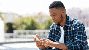 Man of color sitting outdoors on a sunny day, looking at his phone.