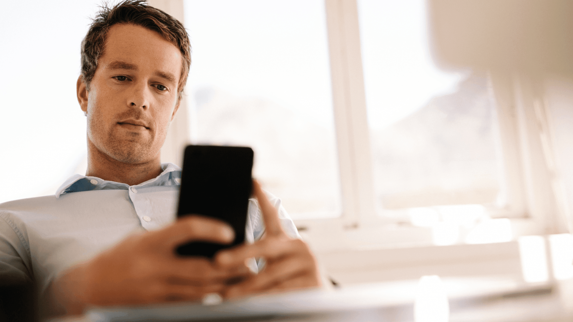 Man sitting and using his phone, with a serious expression while engaged in conversation.