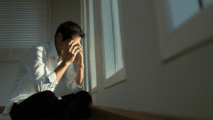 Man sitting on the floor by a window, crying with his hand covering his face.