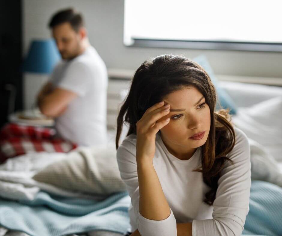A fed up woman sits on the end of the bed while a man turns his back on her and folds his arms