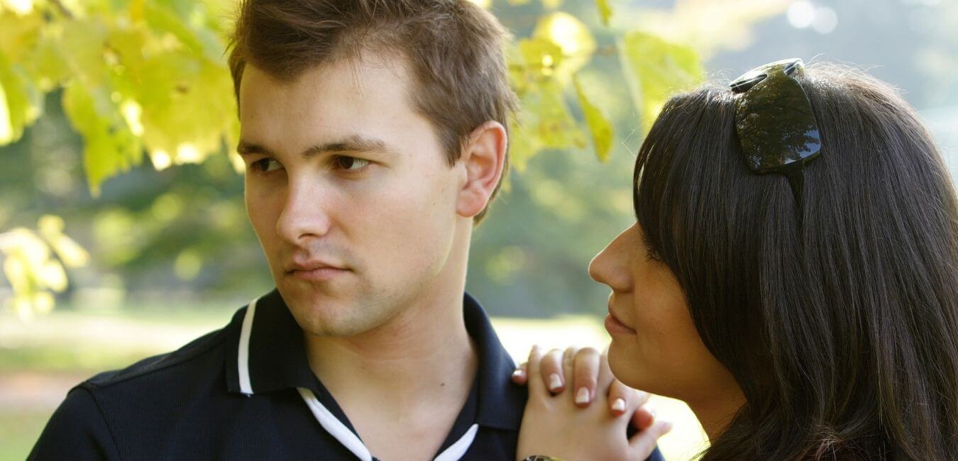 A man looking away looking disinterested while a woman leans on his should looking at him with love