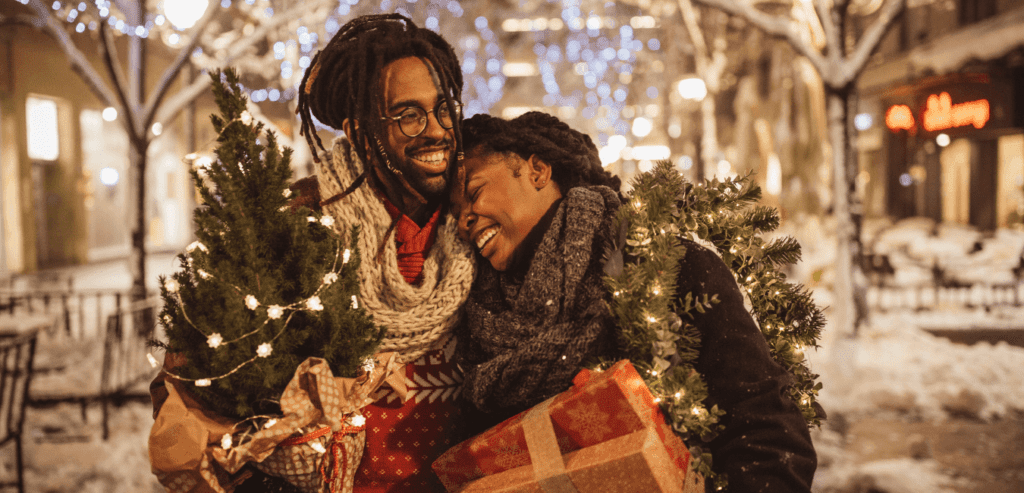 A young black couple stand close together she has her head on his shoulder, she is holding gifts and he has a small christmas tree, they are very happy