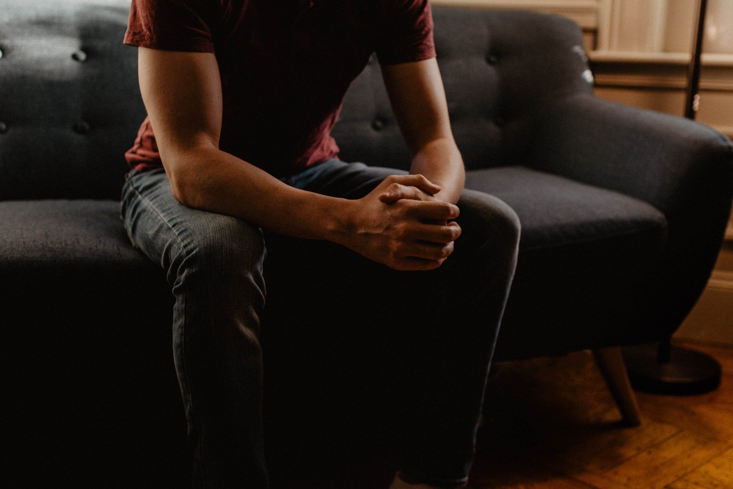 Man wearing red shirt sitting on couch holding his hands together in a room