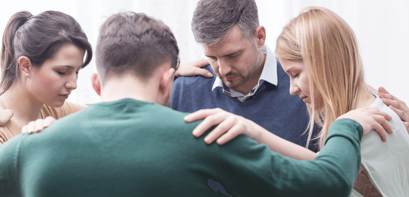 Christian people holding each others elbow bowing head in a room for group therapy session