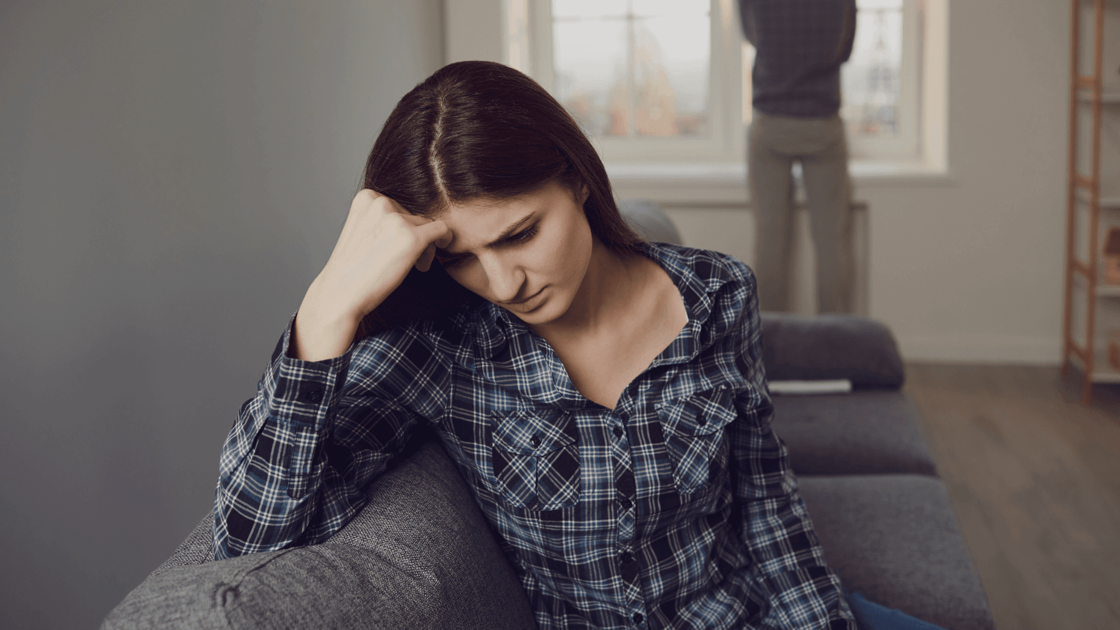 At home, woman sits on couch, sad while man stands at window, distant