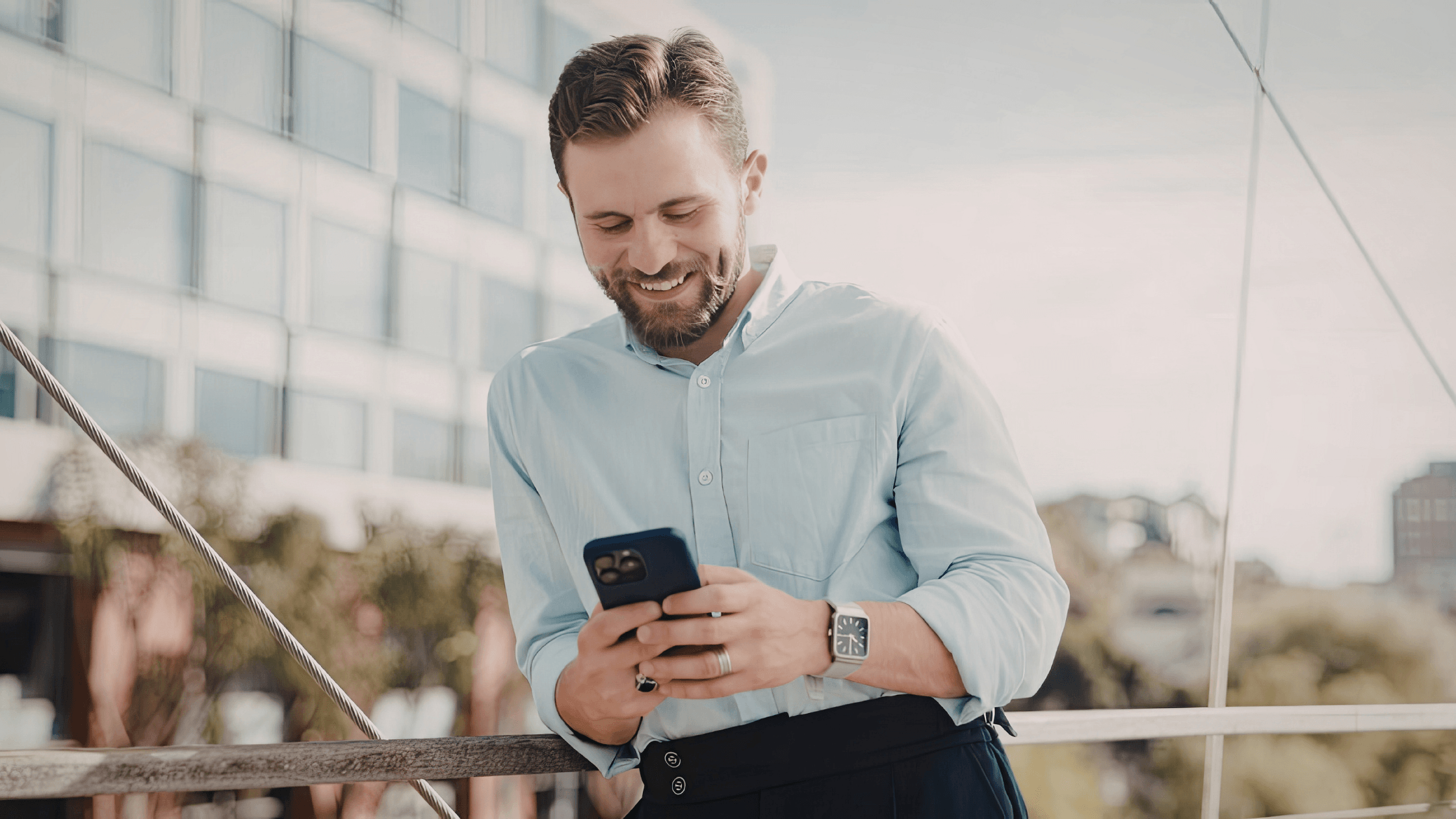 Man in formal clothes stands outdoors, smiling at his phone.