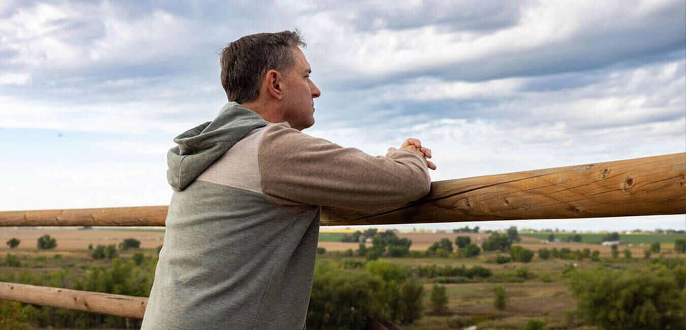 A man standing outdoors leaning on a wooden railing, looking out over a wide landscape, reflecting a moment of thought, clarity, and personal reflection.