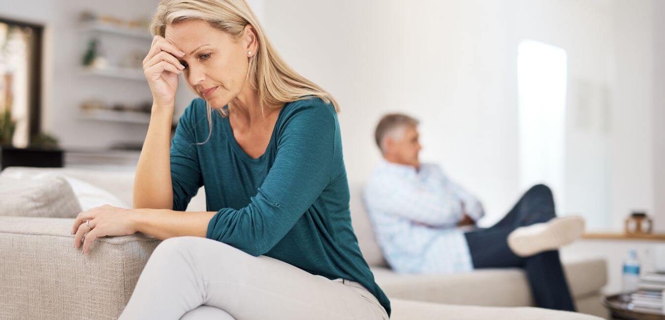 Couple sitting on a couch at home after an argument, woman visibly upset in the foreground while the man sits in the background.