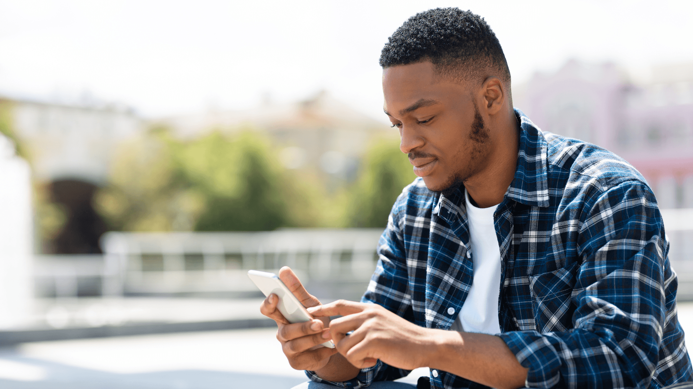 Man of color sitting outdoors on a sunny day, looking at his phone.