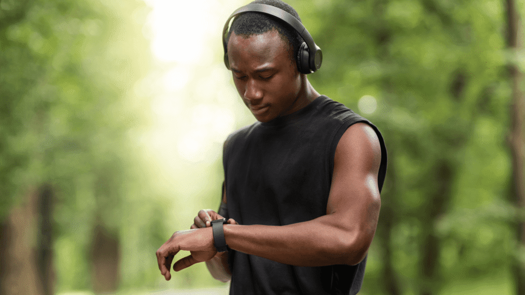 Man of color outdoors in headphones, looking at his smartwatch before running.