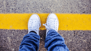 Close-up of a person in jeans and white sneakers standing on a yellow road line marking the boundary.