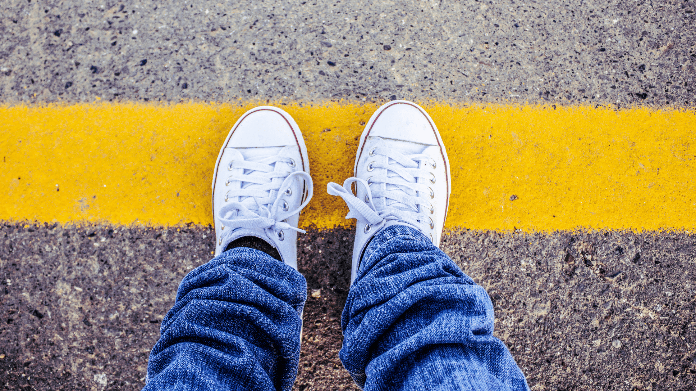 Close-up of a person in jeans and white sneakers standing on a yellow road line marking the boundary.