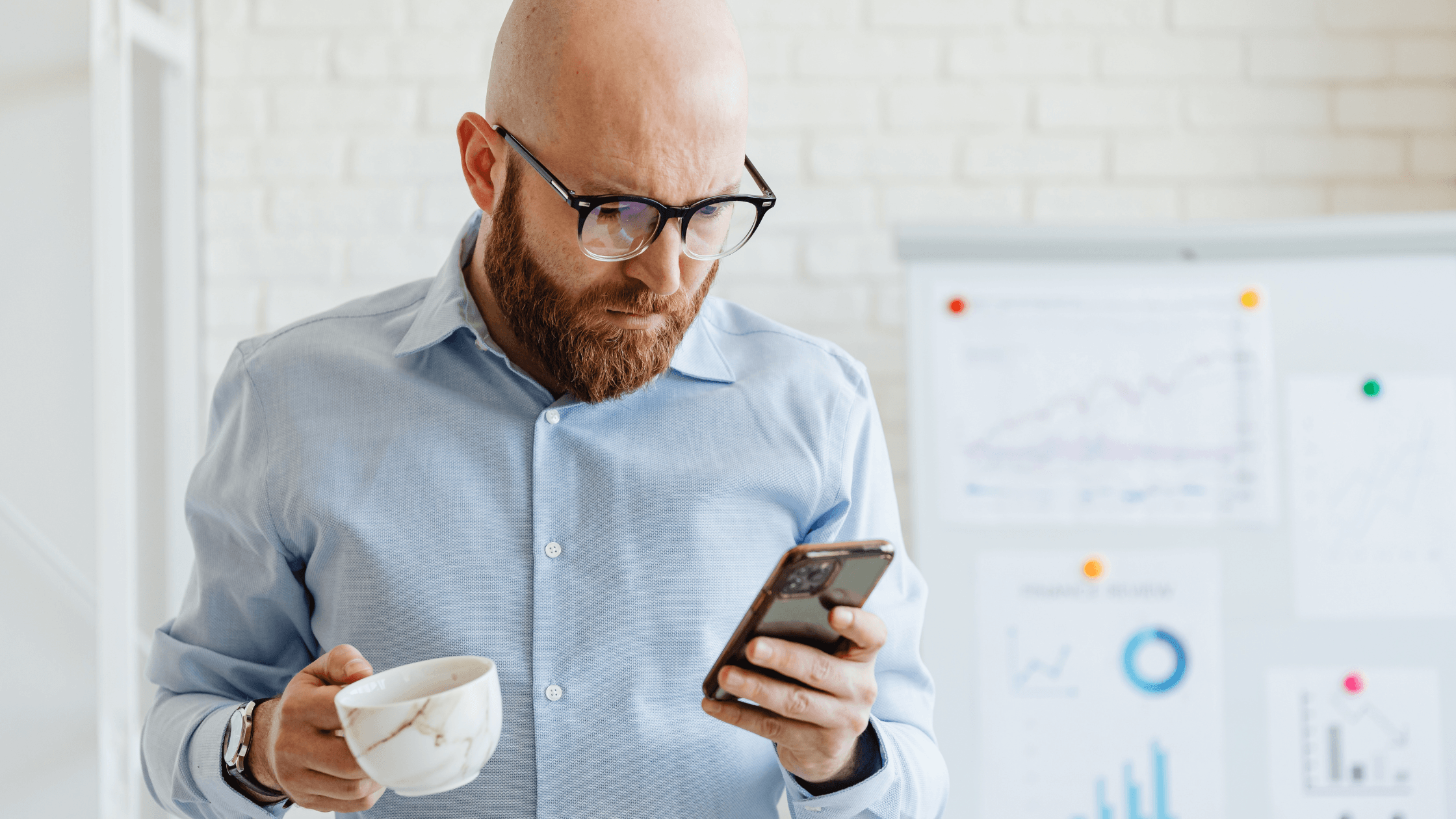 Man in an office holding a cup in one hand and a phone in the other, appearing busy on his phone.