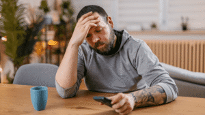 Man seated at a table with one hand on his head and the other holding a phone, appearing troubled.