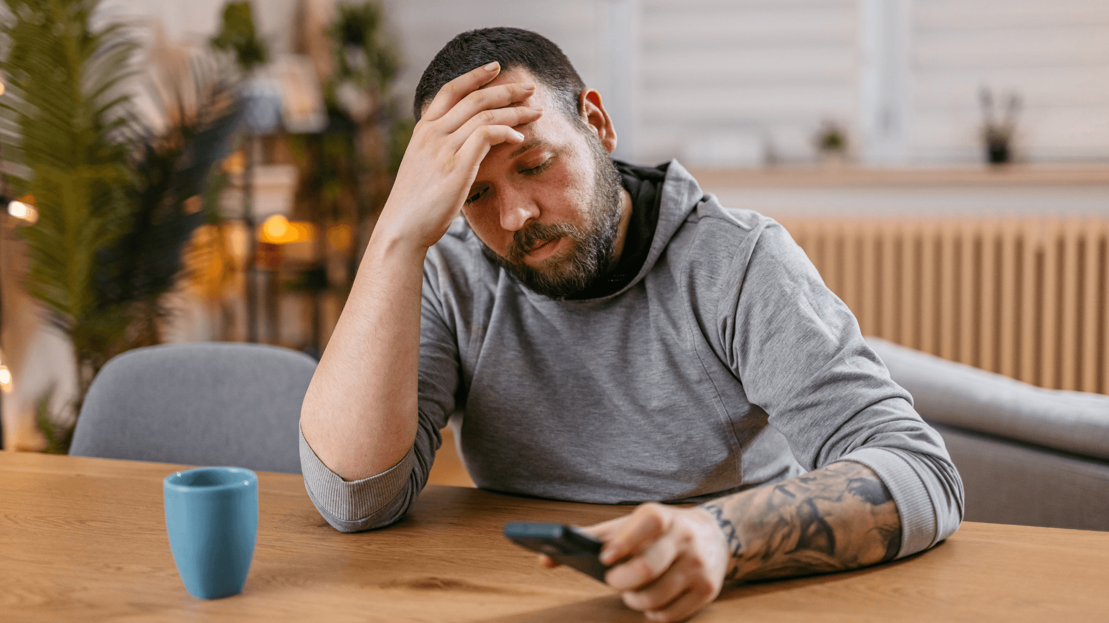 Man seated at a table with one hand on his head and the other holding a phone, appearing troubled.