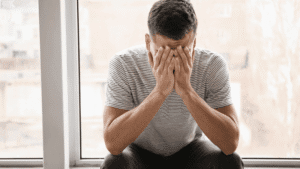 Man alone at home near windows, looking unhappy with his head lowered and hand on his face.