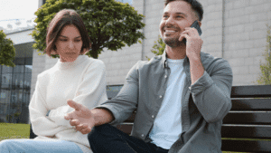 Couple seated outdoors, the man talking on his mobile phone while the woman looks unhappy.
