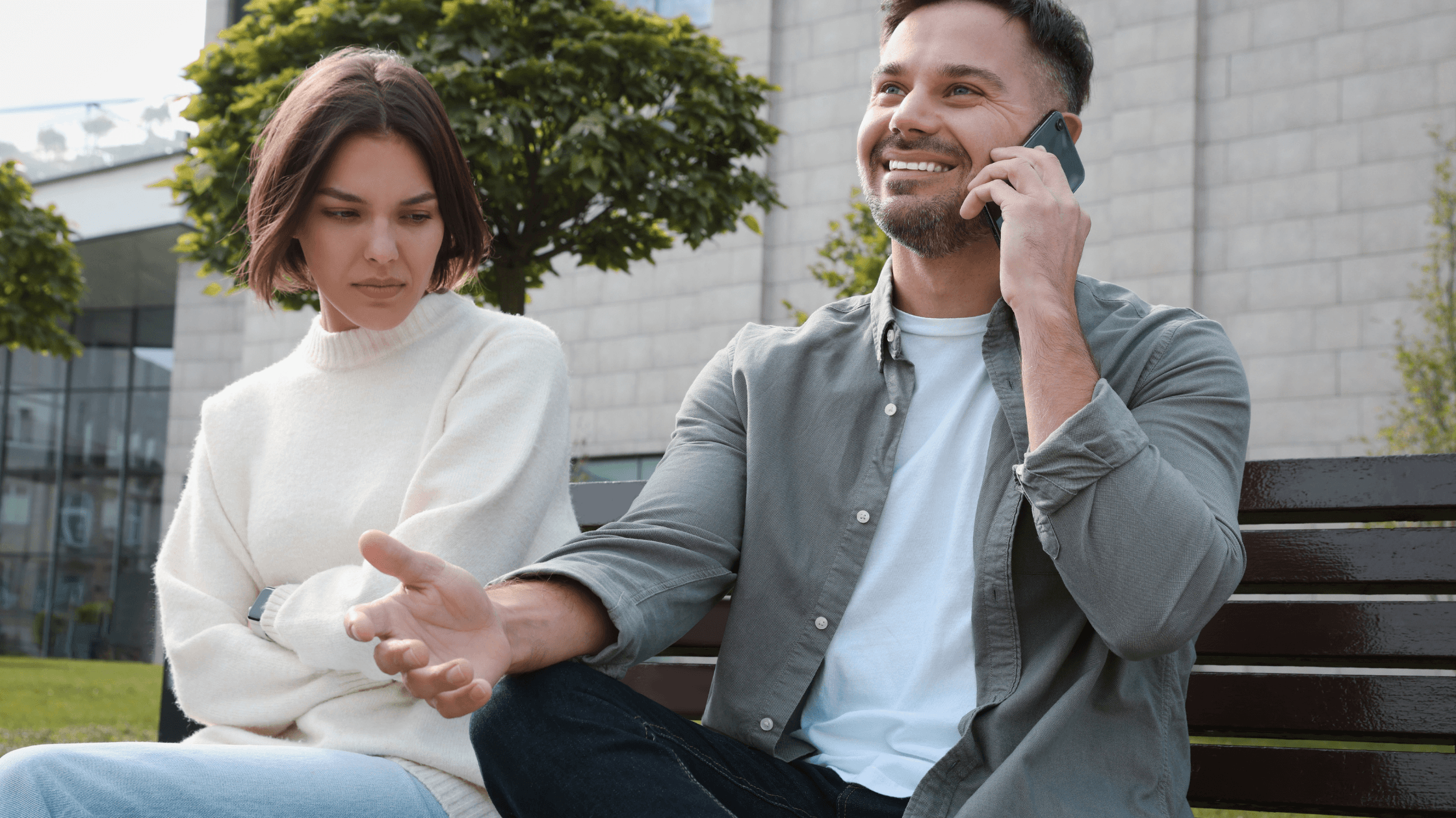 Couple seated outdoors, the man talking on his mobile phone while the woman looks unhappy.