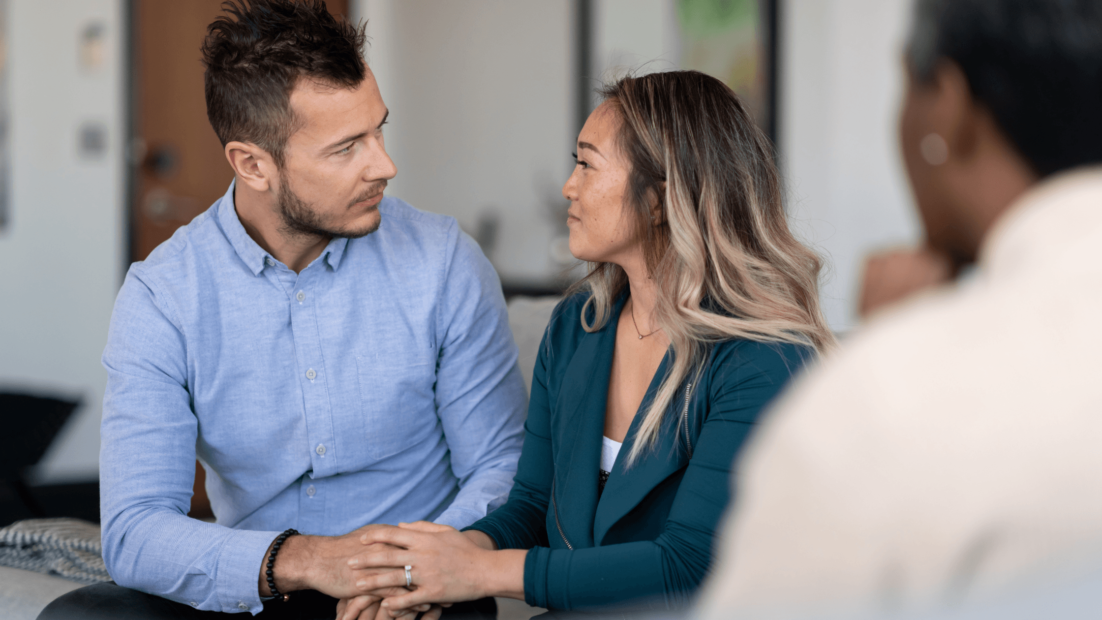 Couple seated together, holding hands and looking at each other while speaking with a counselor.