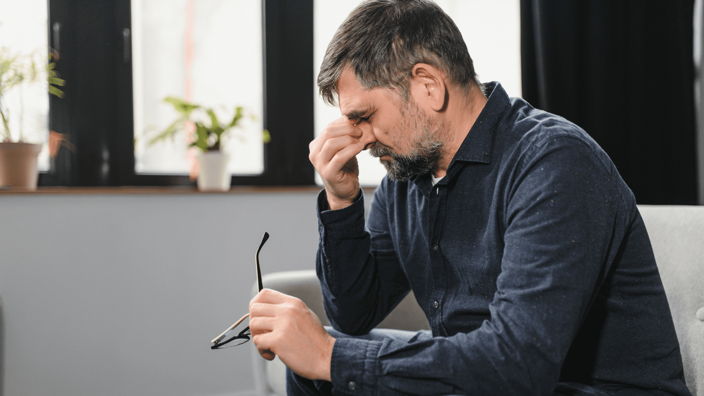 Man sitting on a couch at home, one hand on his nose and the other holding his glasses, appearing to cry.