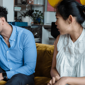 A man and woman sit on a couch and are looking upset, he is holding his head in one hand