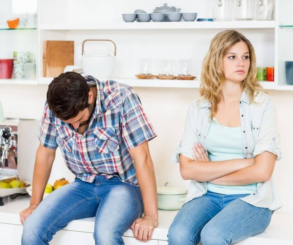 A couple sitting on the kitchen counter. The man is looking down and the woman is folding her arms looking away