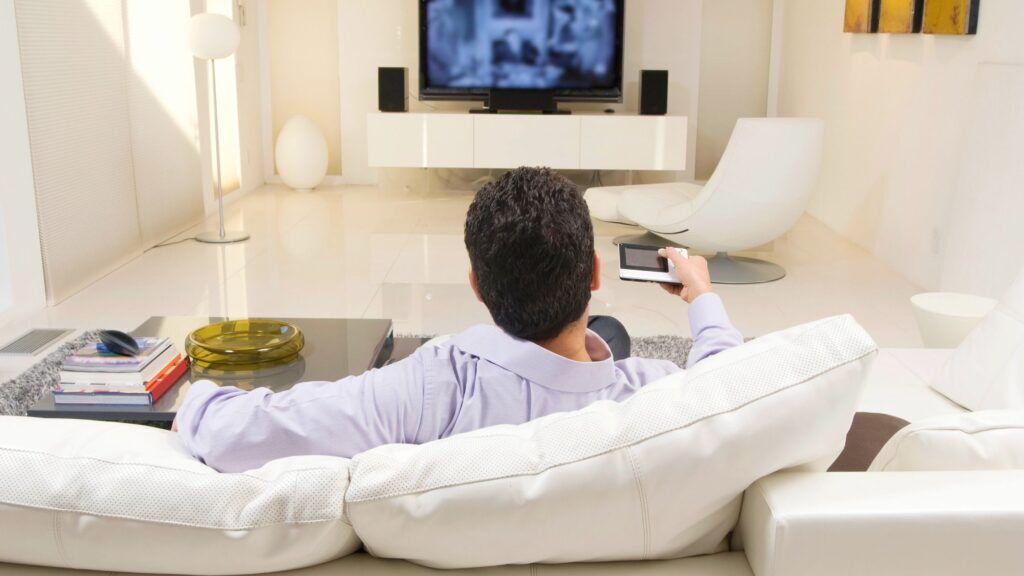 A man sits on a white leather couch, his back faces the camera, he is holding a tv remote and it is pointed at the television