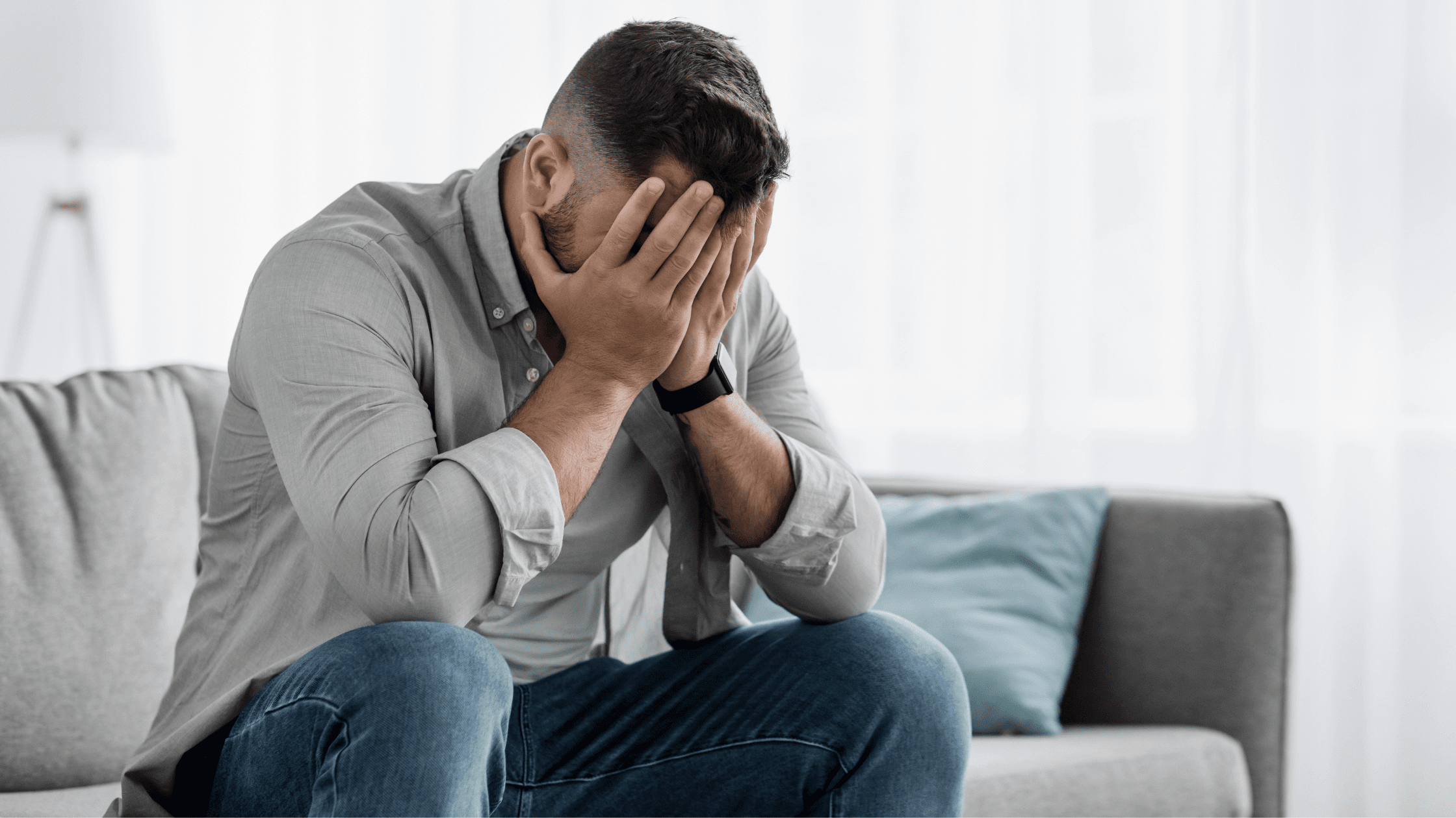 Man sits in silence on his couch, head lowered, hand on his face, battling feelings of shame and addiction.