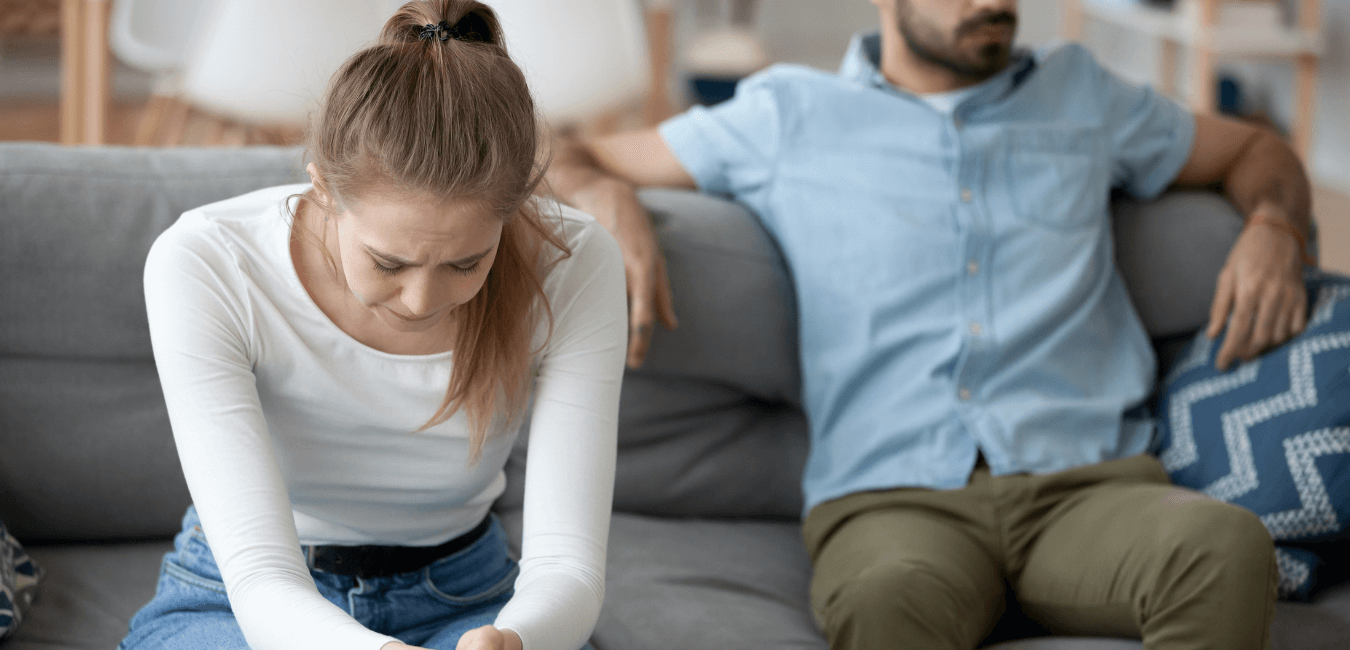 a white heterosexual couple sit on a couch. The woman is distressed and looking down, the male is leaning back on the couch looking away