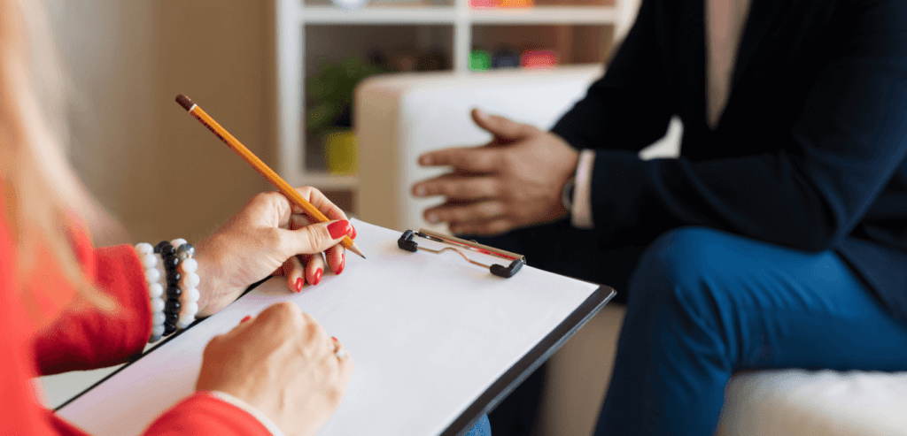 a woman sits in the foreground of the photo, her face is not visible, she holds a clipboard and pencil and is wearing a red sweater. She is talking to a man who is on a couch and his face is not visible.